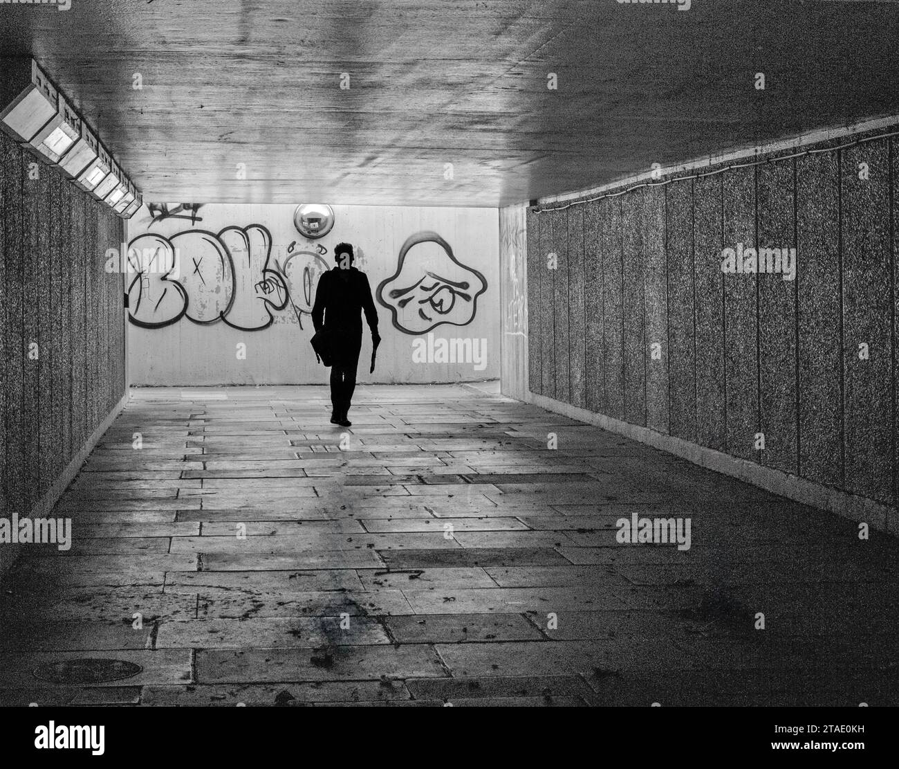 Schwarz-weißes Bild der Silhouette einer Person, die in einer dunklen Fußgängerunterführung läuft, mit Graffiti an der Wand hinter sich. Stockfoto