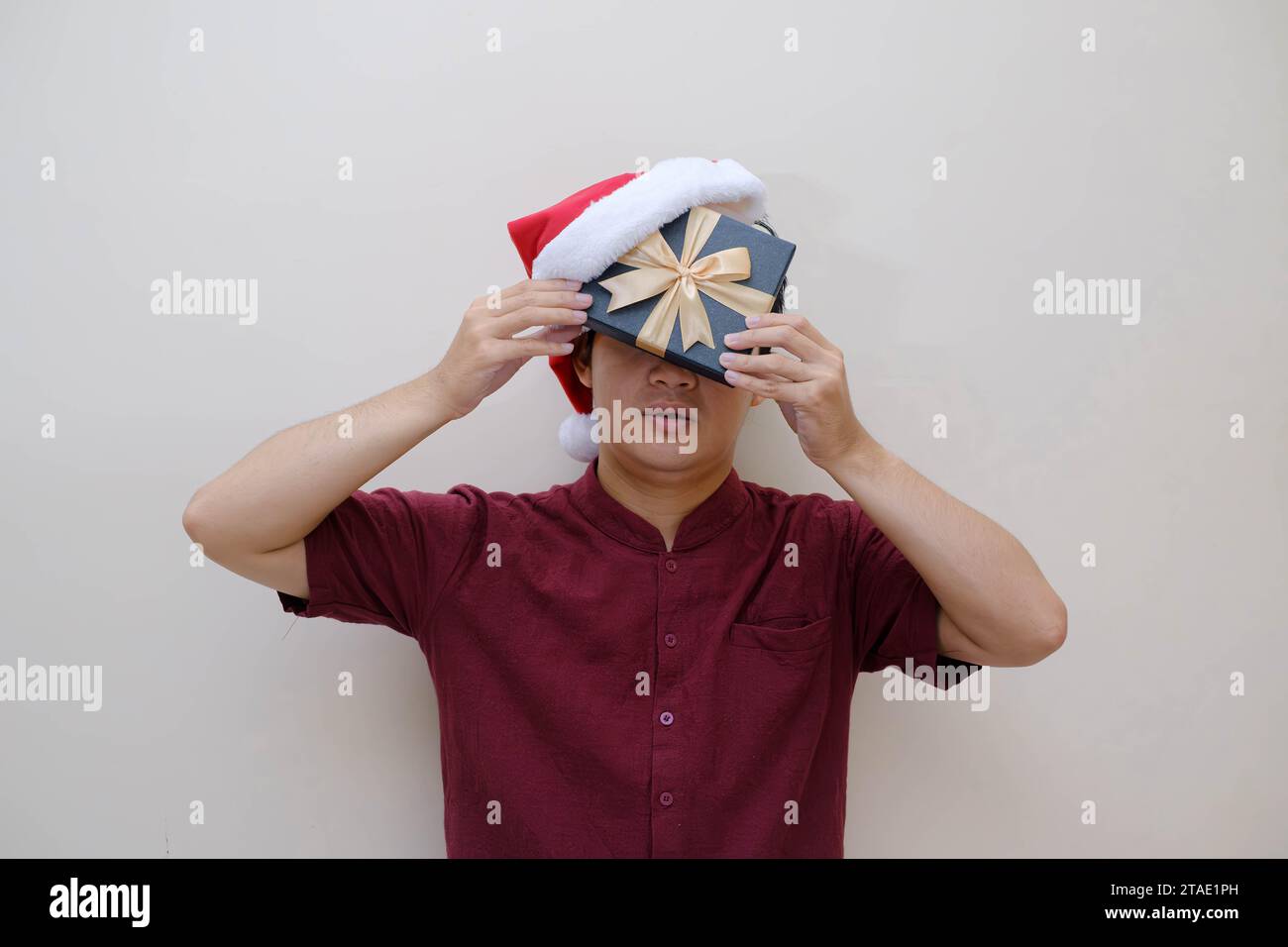 Ein junger asiatischer Mann hält eine Geschenkbox und einen Weihnachtsmann-Hut in der Hand, die sein Gesicht zur Hälfte bedeckten. Isolierter beigefarbener Hintergrund. Konzeptionelles Weihnachtsfoto. Stockfoto