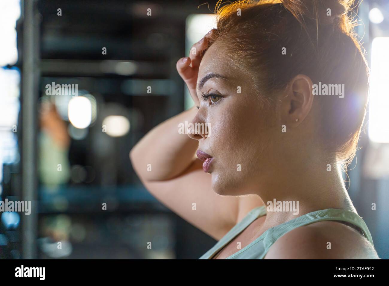 Eine auf Fitness fokussierte Frau nimmt sich während ihres Workouts einen Moment Zeit, um sich auszuruhen. Die Ambient Gym Lampen betonen ihren kontemplativen Ausdruck und zeigen ein an Stockfoto