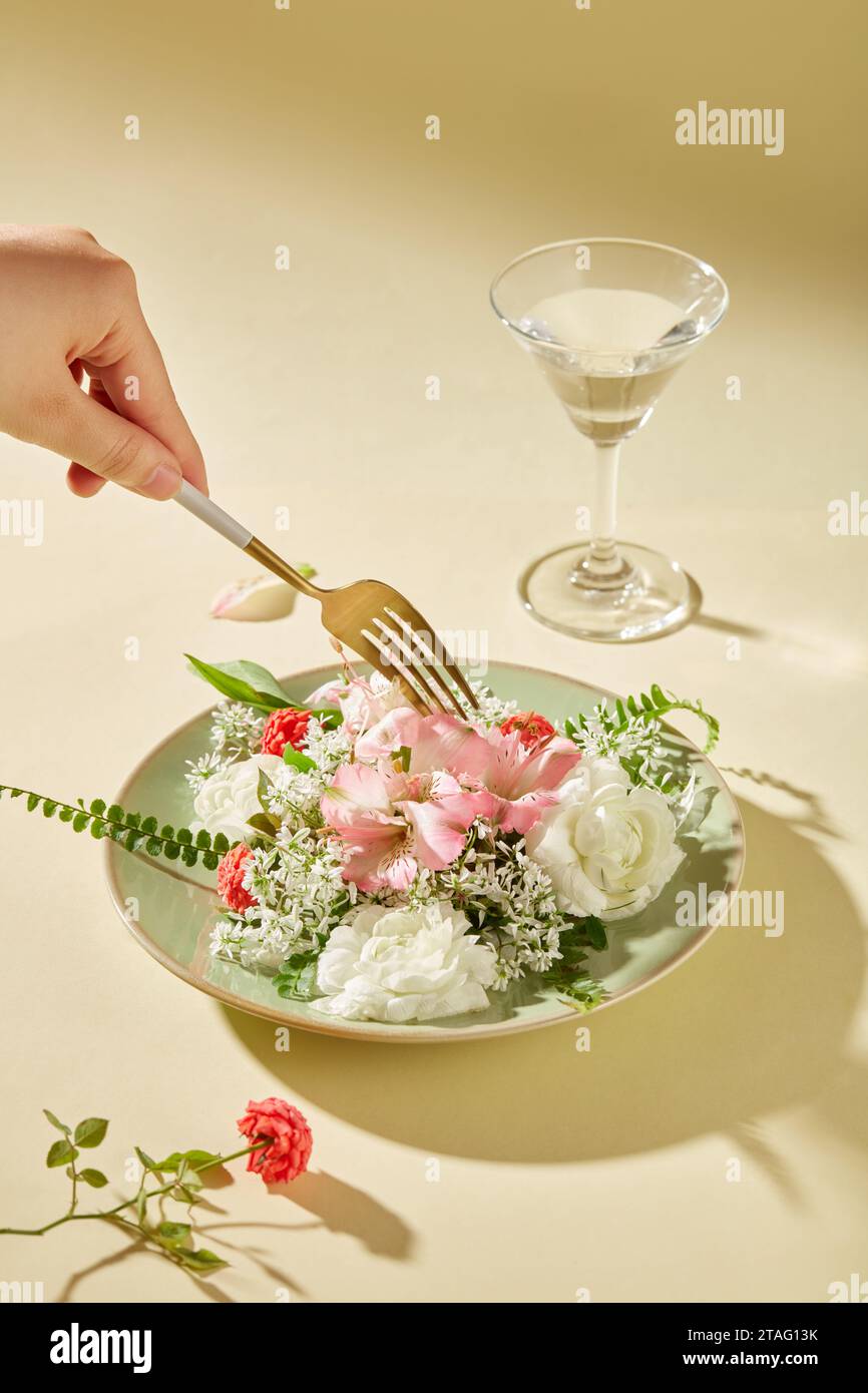 Die Hand der Frau hält eine goldene Metallgabel, auf einer Keramikplatte stehen verschiedene Blumen neben einem mit Wasser gefüllten Cocktailglas. Künstlerisch Stockfoto
