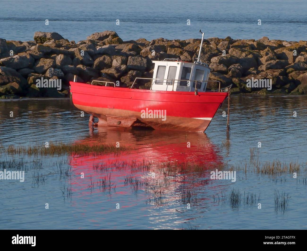 Bei Ebbe wird ein kleines rot-weißes Boot in der Nähe von Felsen vor Anker gebracht. Er kippt um und wird im Flachwasser reflektiert. Stockfoto
