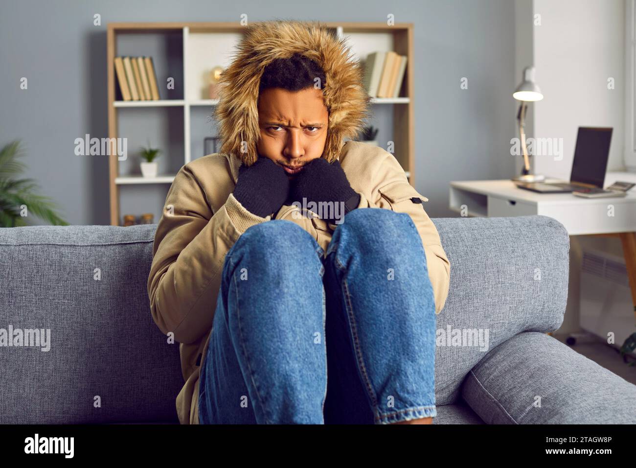 Gefrorener afroamerikaner sitzt in einem Wintermantel auf dem Sofa zu Hause. Heizungsprobleme Konzept. Stockfoto