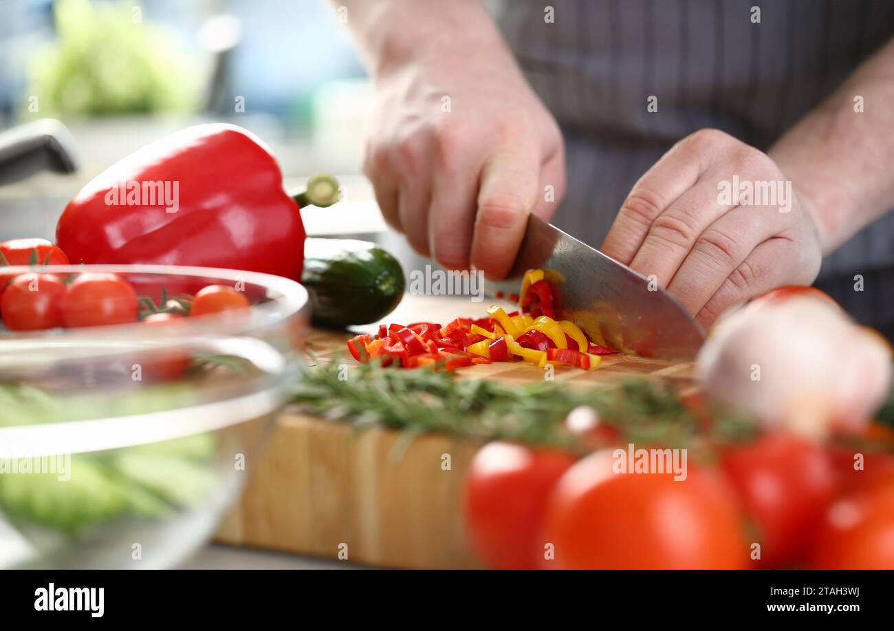 Die Hände des Kochs schneiden gelbe und rote Paprika auf dem Schneidebrett zusammen mit anderem Gemüse auf dem Tisch in der Küche Stockfoto