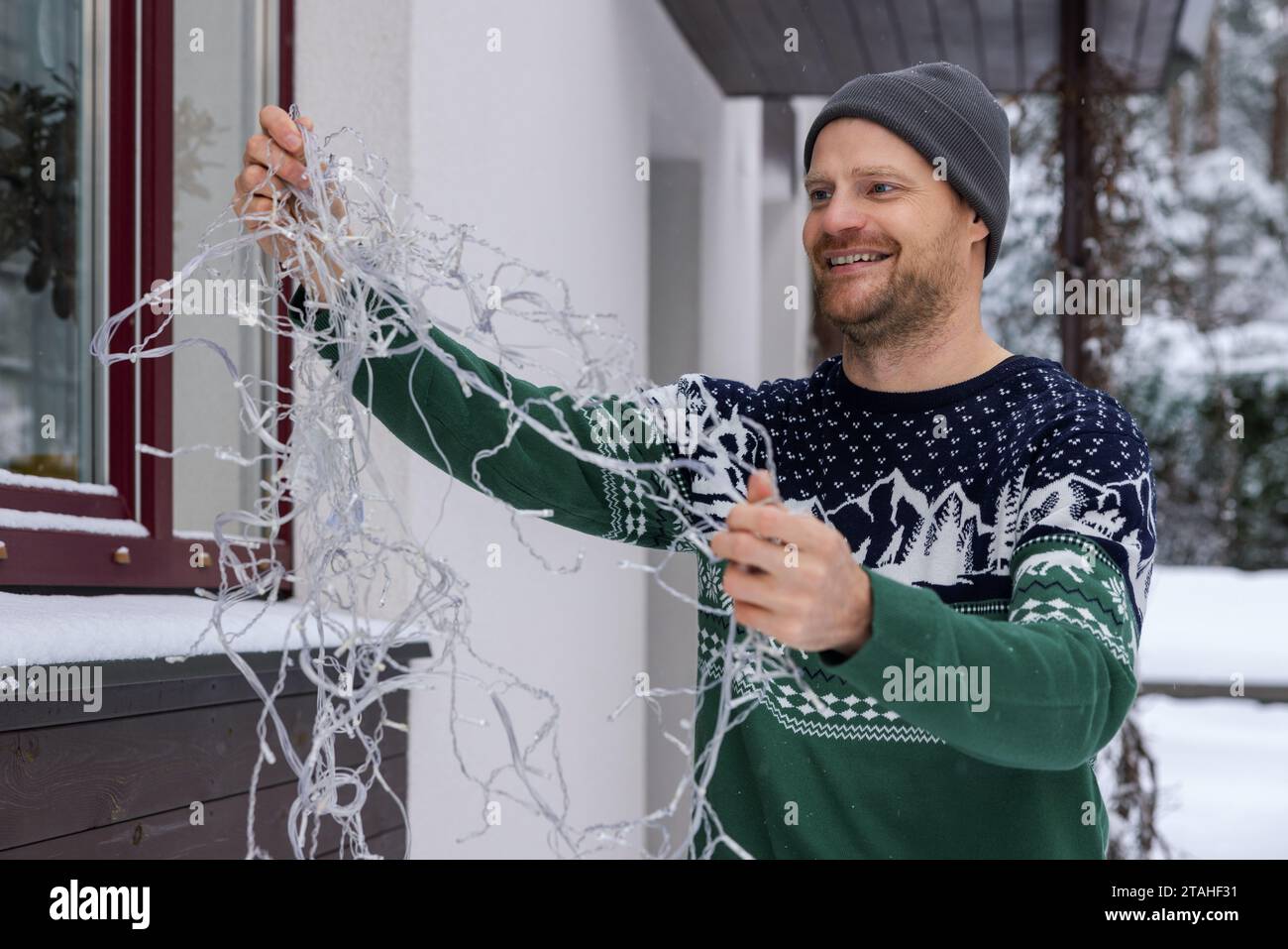 Glücklicher Mann draußen im weihnachtspullover, der Lichterketten für die Außendekoration des Hauses entwirrt Stockfoto