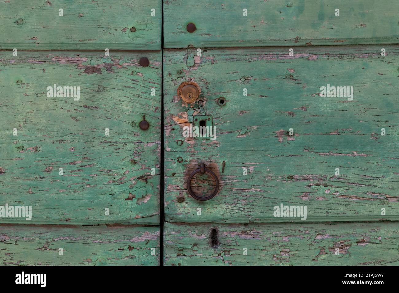 Eine verblasste, abblätternde und verwitterte, hellgrün lackierte Tür mit abgebrochener Oberfläche, Schlüssellöchern, Schloss und rostfreiem Metallklopfer. Stockfoto