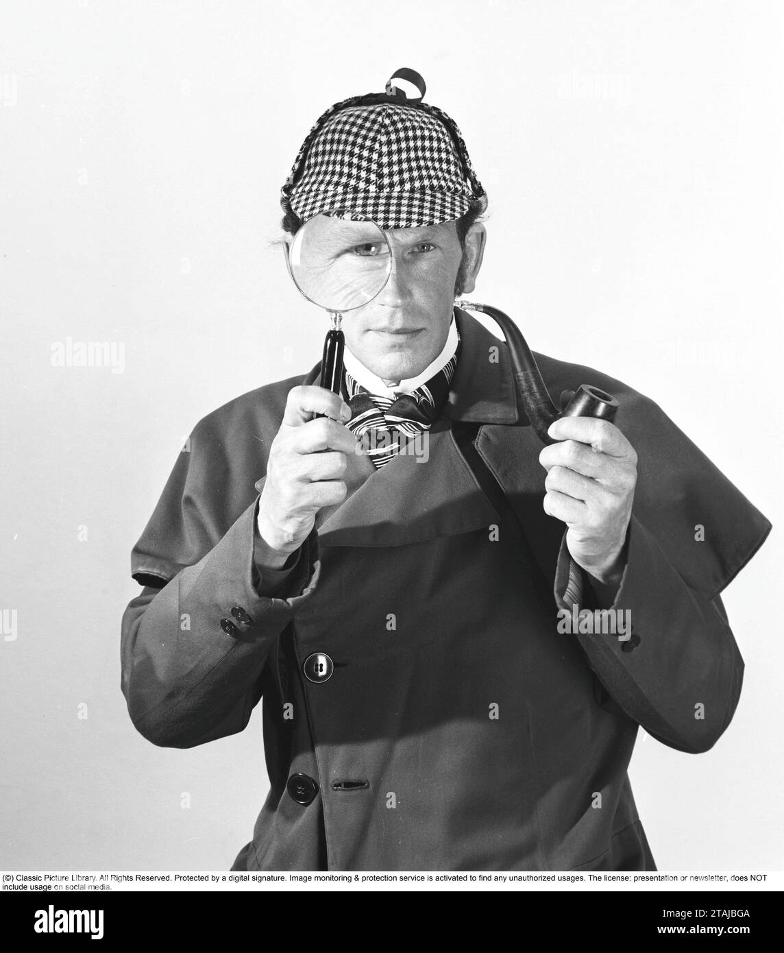 Detective spielen. Ein Mann in den typischen Kleidern des berühmten fiktiven Detektivs Sherlock Holmes schaut durch eine Lupe in die Kamera. Stockfoto