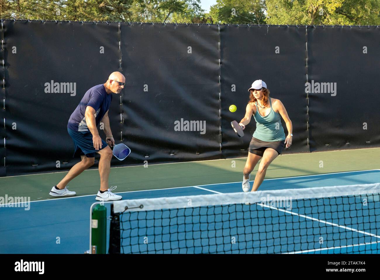 Ein weiblicher Pickleball-Spieler gibt eine Fahrt in der Nähe der Baseline zurück, während ihr Partner sich auf das Netz zubewegt. Stockfoto
