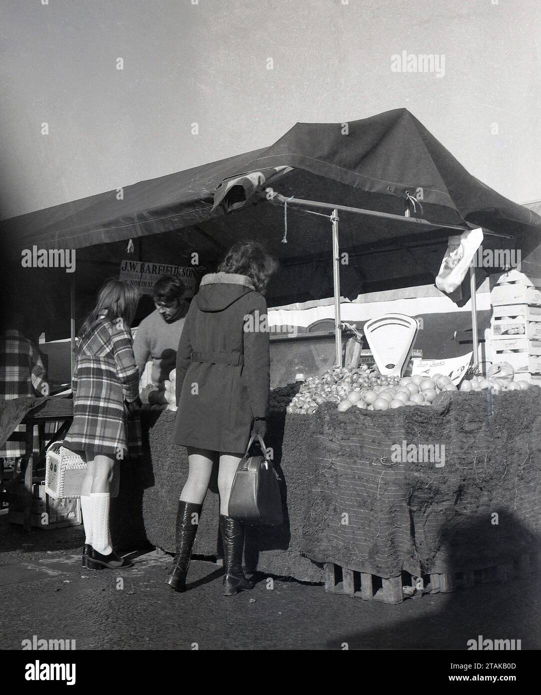 1970er Jahre, historisch, auf einem Bauernmarkt, zwei weibliche Käuferinnen an einem Obst- und Gemüsestand von J. W. Barfield & Son, England, Großbritannien. Die Märkte der Landwirte ermöglichen es den Lebensmittelerzeugern und lokalen ländlichen Unternehmen, ihre Erzeugnisse direkt an die Öffentlichkeit zu verkaufen. Stockfoto