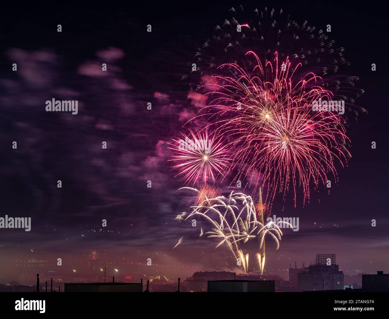 Rotes Feuerwerk brach am Mitternachthimmel. Festliche Feuerwerke über der kleinen Stadt, die Feiertage feiern. Pyrotechnisches und Feuerwerkskörper-Konzept. Stockfoto
