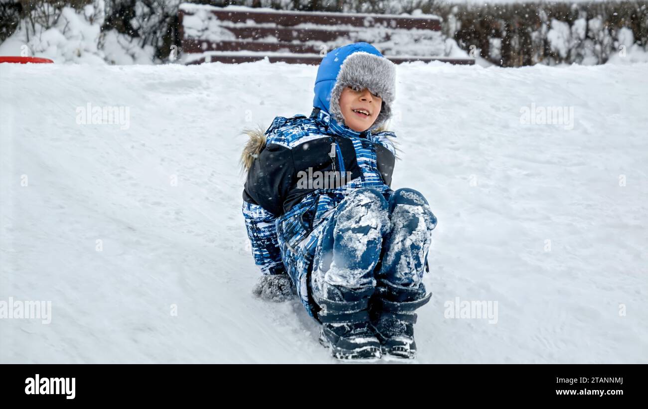 Die Freude eines Kindes, wenn es einen schneebedeckten Hügel mit den Füßen hinunterfährt, wobei das Filmmaterial verlangsamt wird, um jeden Moment der aufregenden Fahrt zu betonen Stockfoto