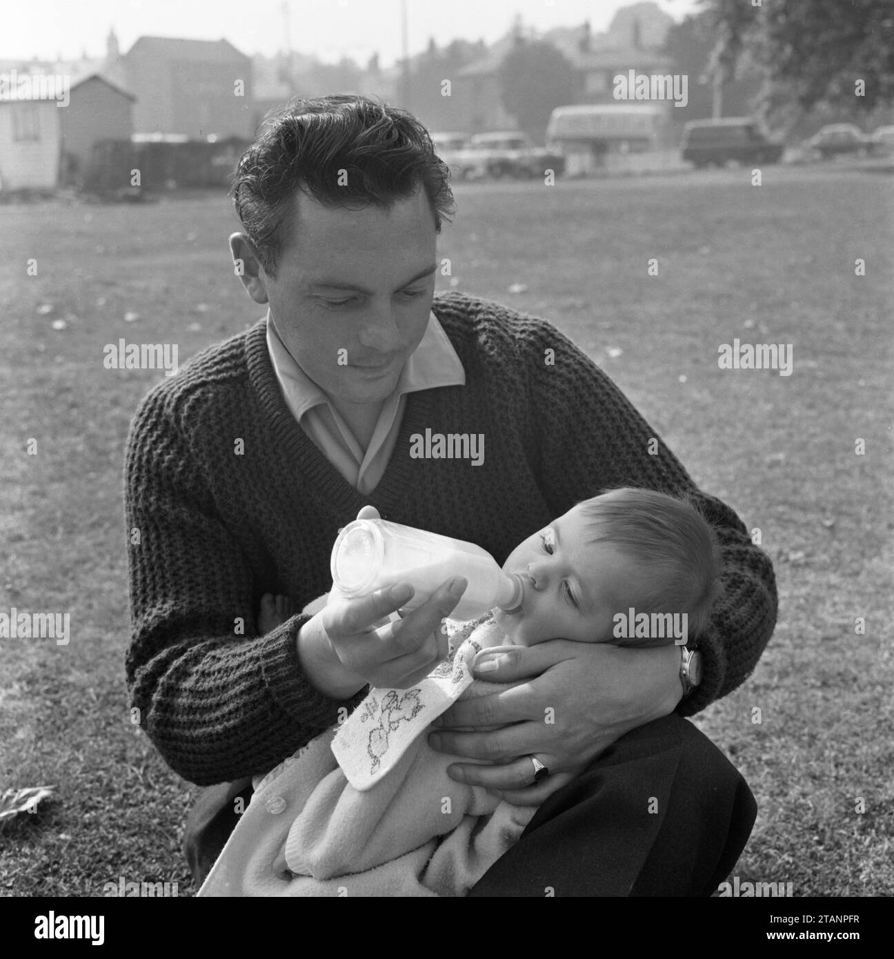 Eine Vaterflasche aus den 1960ern, die einem kleinen Baby Milch in einem Vorstadtpark füttert Stockfoto