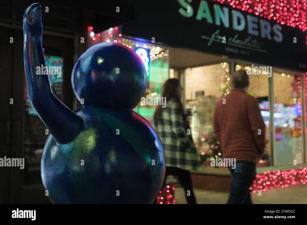 Skulptur einer Figur, die vor einem festlichen Geschäft winkt, mit Weihnachtslichtern bei Nacht Stockfoto