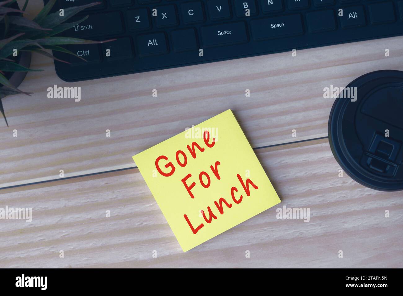 Gelbe Haftnotiz mit Text „Gone for Lunch“ auf einem Holztisch mit Computertastatur, Topfplan und Einwegkaffeetasse. Stockfoto