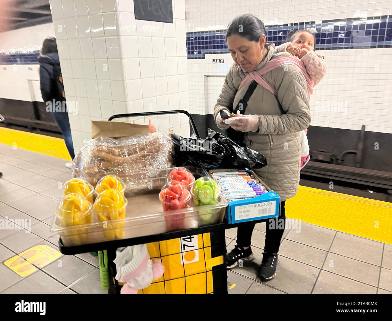 Eine eingewanderte hispanische Frau verkauft Obstbecher mit einem Baby auf dem Rücken auf einem U-Bahn-Bahnsteig in Manhattan, New York City. Stockfoto