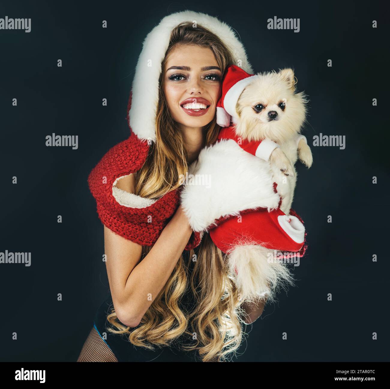 Lächelnde Weihnachtsfrau mit langen Haaren im Pelzhut und weißen Handschuhen umarmt Pommersche Spitze in Weihnachtskleidung und Weihnachtsmann-Hut. Tiere im Urlaubsstil Stockfoto