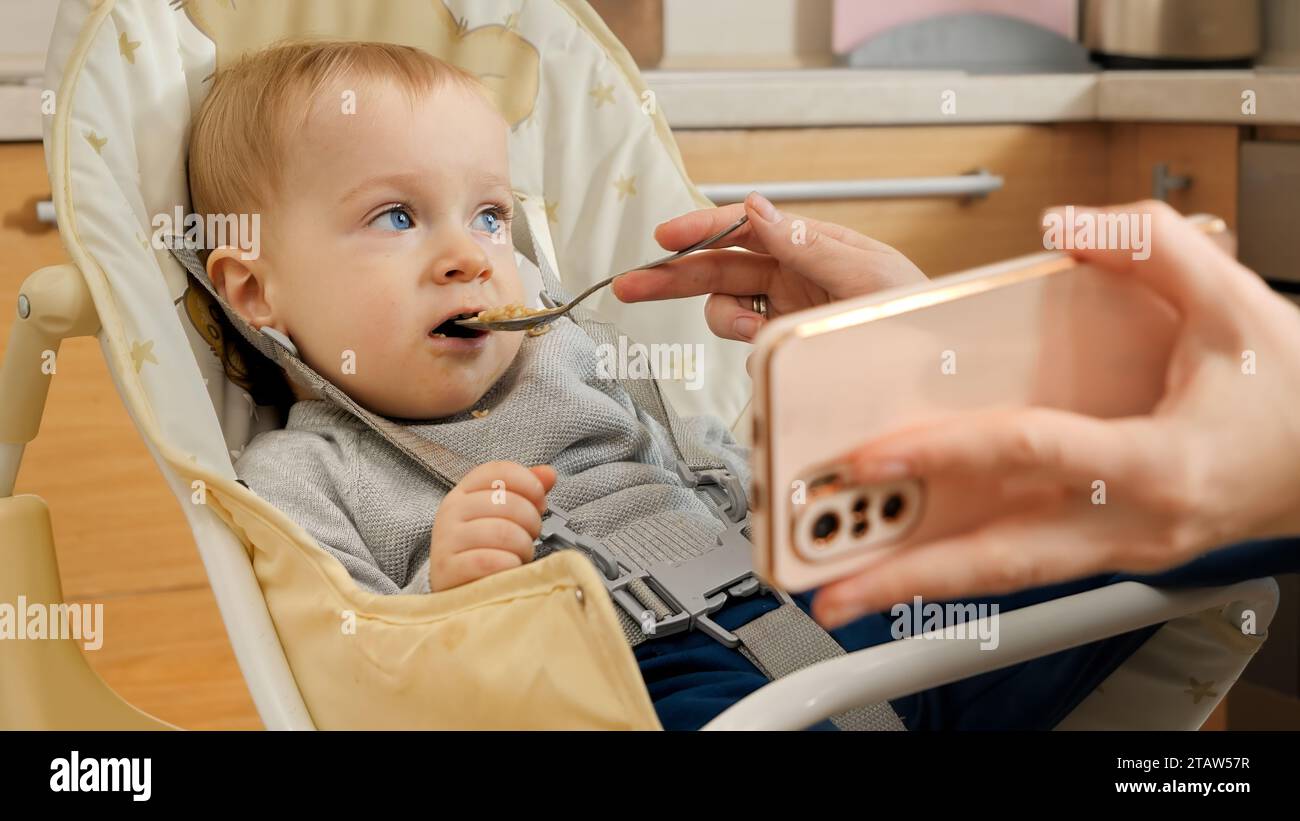 Kleiner Junge, der während des Frühstücks im Hochstuhl Videos auf dem Smartphone ansieht. Konzept der Erziehung, der gesunden Ernährung und der Säuglingsernährung Stockfoto