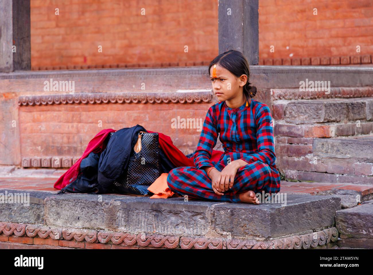 Ein heimisches, dunkelhaariges, hinduistisches Mädchen mit einer orangen Tilaka-Markierung auf der Stirn sitzt im Kreuz im Pashupatinath-Tempel in Kathmandu, Nepal Stockfoto