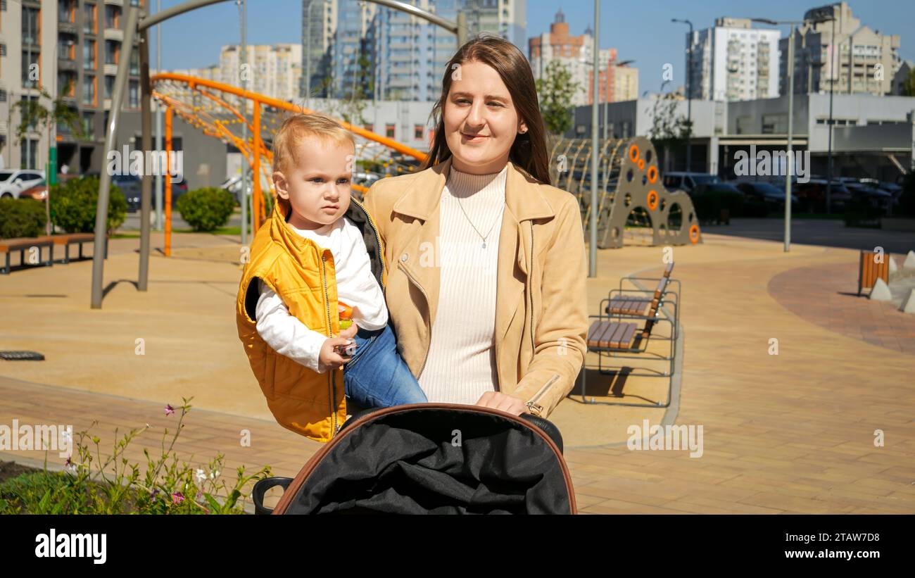 Schöne Mutter hält ihren kleinen Sohn fest, der auf der Straße läuft und den Kinderwagen schiebt. Glückliche Eltern, Familie, die Zeit zusammen hat, Kinder und Eltern Stockfoto