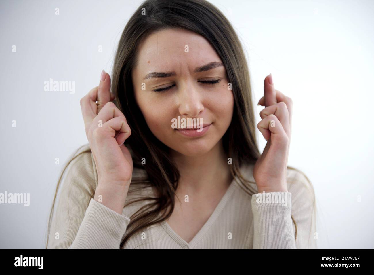 Zufrieden stellende attraktive Europameisterin wünscht sich zu gewinnen, hebt mit gekreuzten Fingern die Hände, wartet auf das Lottoergebnis, schließt die Augen, in modischem Kleid gekleidet, isoliert über der weißen Wand Stockfoto