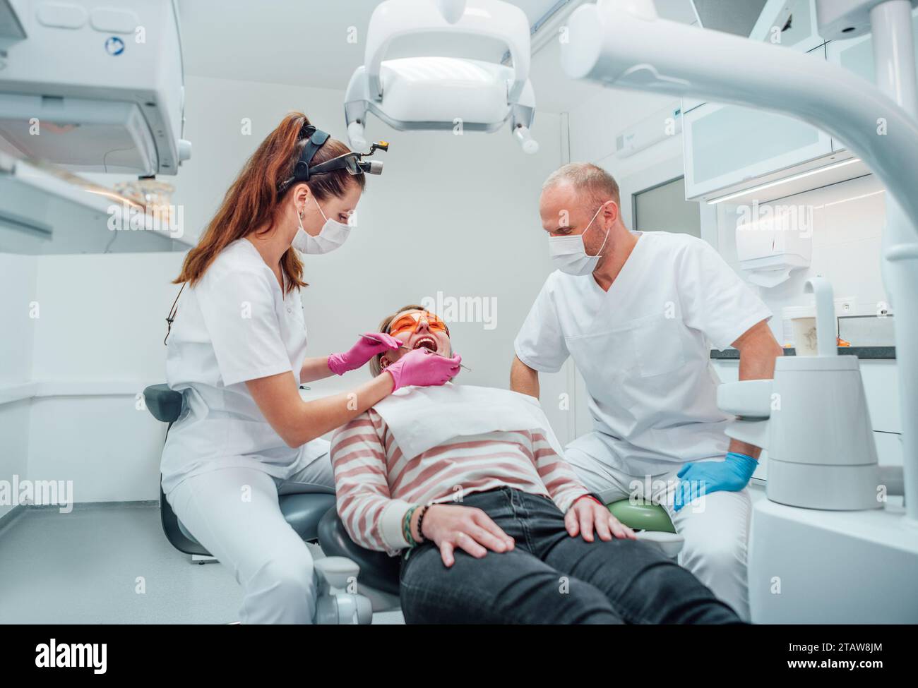 Zahnärztlicher Klinikpatiententermin. Zahnarztfrau in Lupenbrille, die Zahnprophylaxe mit medizinischen Werkzeugen macht. Junger Mann, der ihr hilft. Er Stockfoto