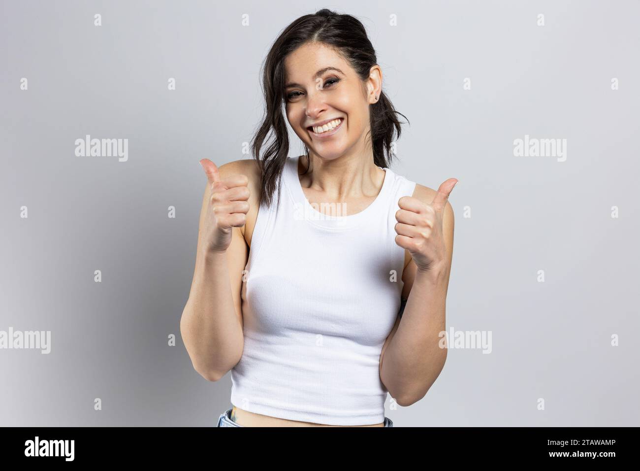 Fröhliche junge Frau macht positives Zeichen mit dem Daumen nach oben, positive Stimmung, erfolgreiche Geste, auf hellgrauem Studiohintergrund Stockfoto