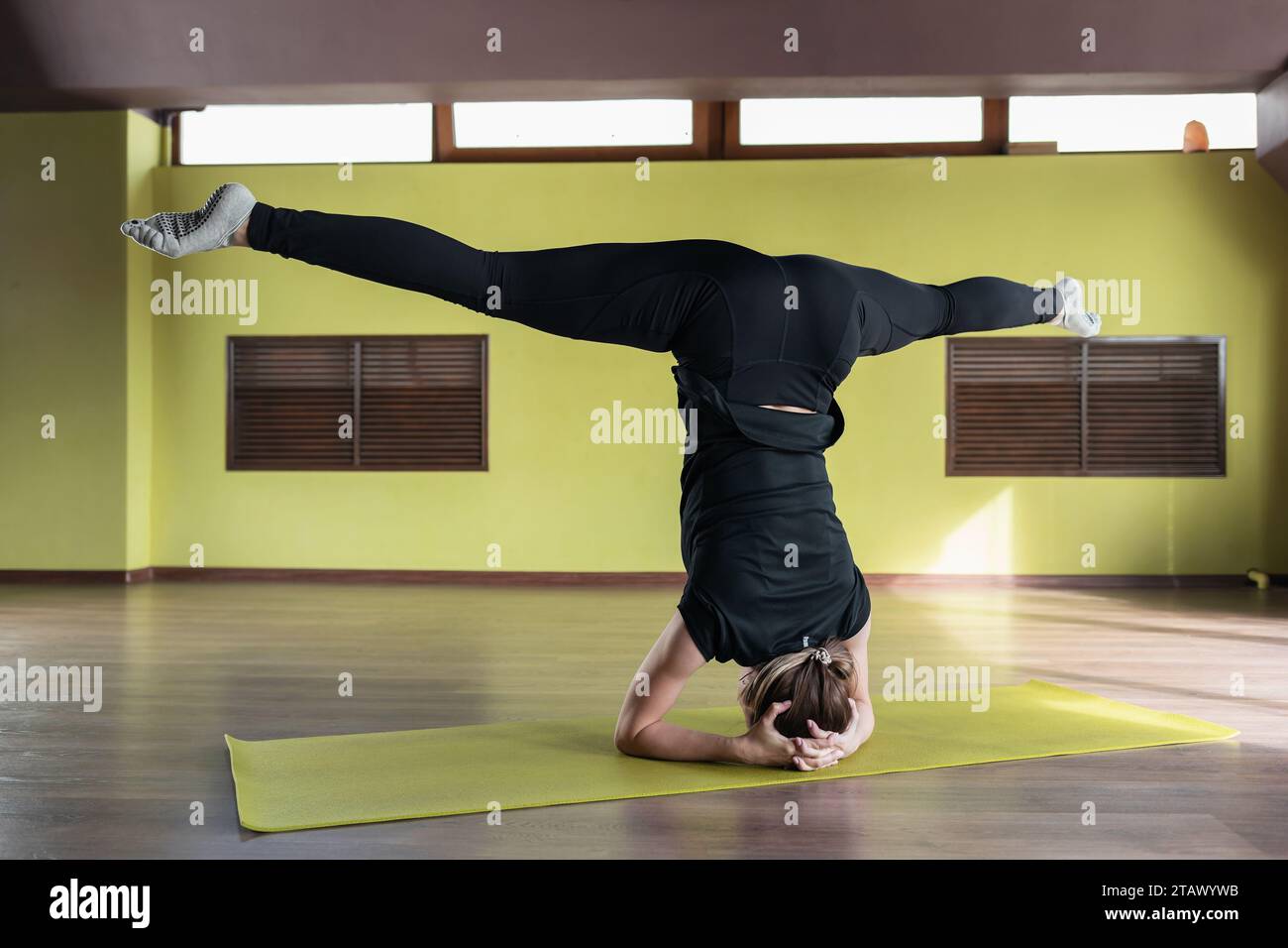 Junge Frau, die Übungen Shirshasana mit Samakonasana durchführt, Kopfstand mit Ellbogen gestützt, Training in schwarzen Leggings und T-Shirt im Studio auf Matte Stockfoto