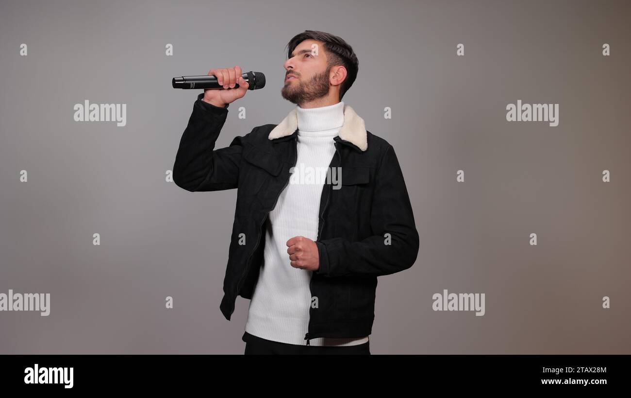 Ein junger Mann, der ein Mikrofon hält und vor grauem Hintergrund singt. Stockfoto