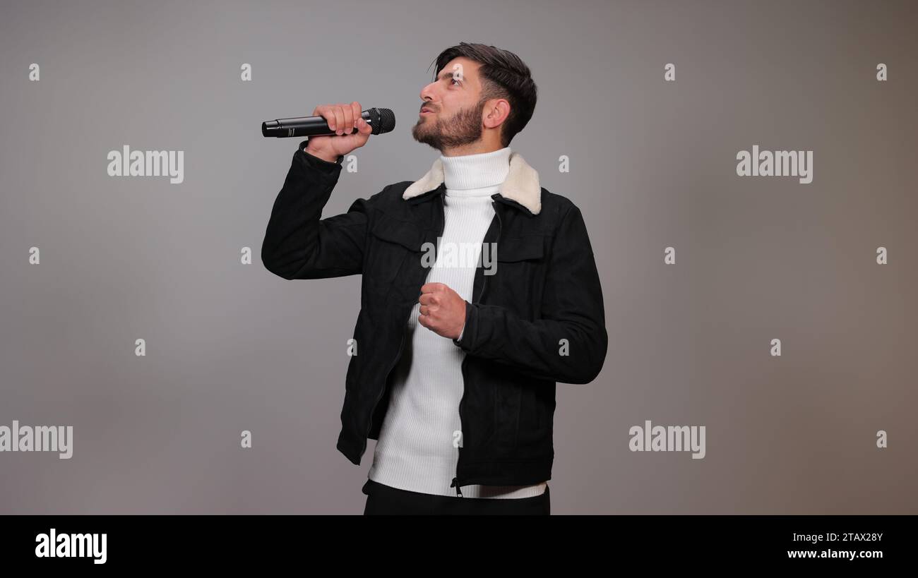 Ein junger Mann, der ein Mikrofon hält und vor grauem Hintergrund singt. Stockfoto
