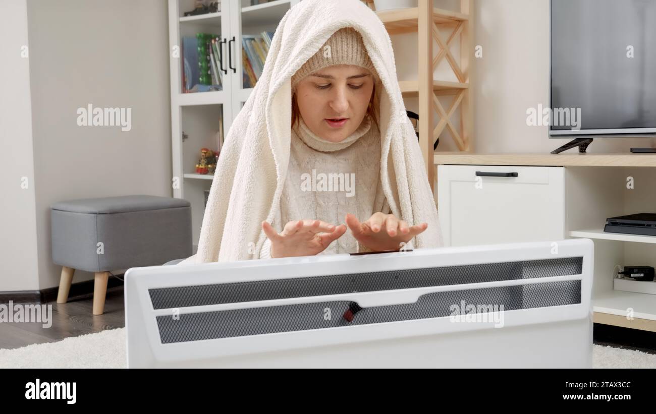 Junge Frau, die sich in Decken-Sitzen neben der elektrischen Heizung bekleidet und die Hände wärmt. Konzept der Energiekrise, hohe Rechnungen, Wirtschaftlichkeit und Einsparung von Geld auf Mo Stockfoto