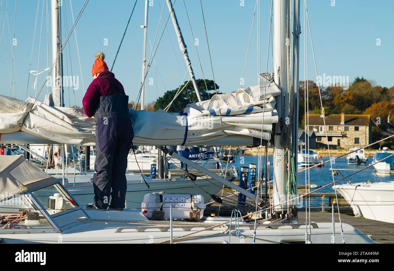 Frau Sailor in warmer Segelkleidung und Wackelhut-Rollen, Abblättern und Binden Eines Hauptsegels auf Einem Segelboot, Yacht, Bembridge Harbour im Winter, Insel Stockfoto