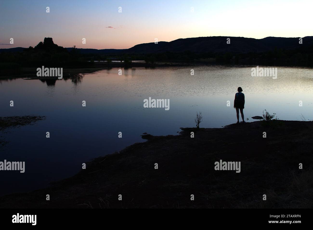 August Abend fällt am Ufer des Salagou-Sees, während eine bewegungslose weibliche Silhouette das dunkle, stille Wasser beobachtet (Hérault, Occitanie, Frankreich) Stockfoto