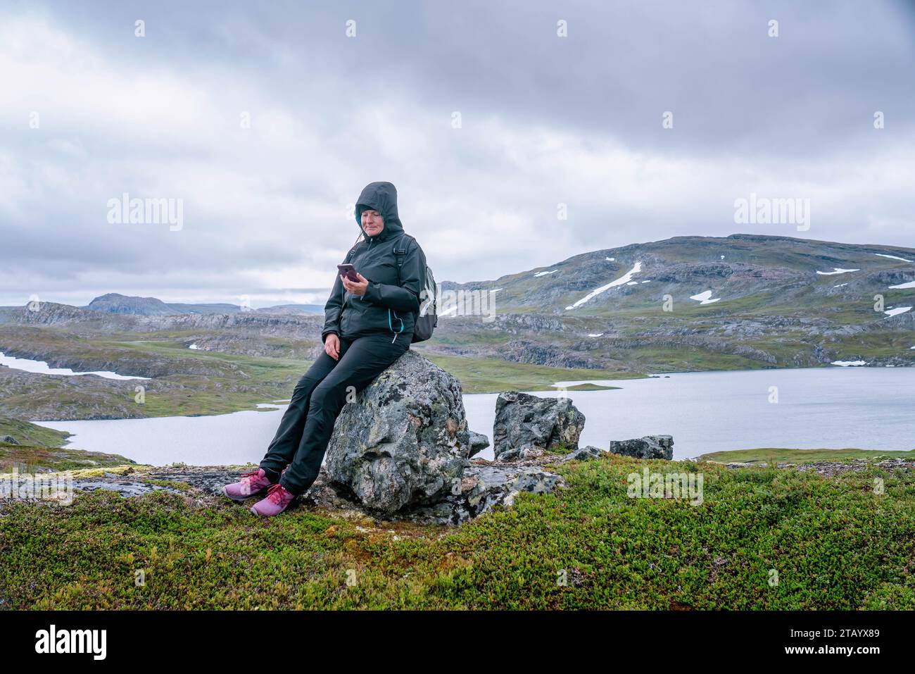 Reife Frau macht eine Pause und ruht sich auf Stein aus, blickt auf ihr Handy und wandert hoch in den norwegischen Bergen. Gesunder Lebensstil. Norwegen, Krutvatnet. Vordere s Stockfoto