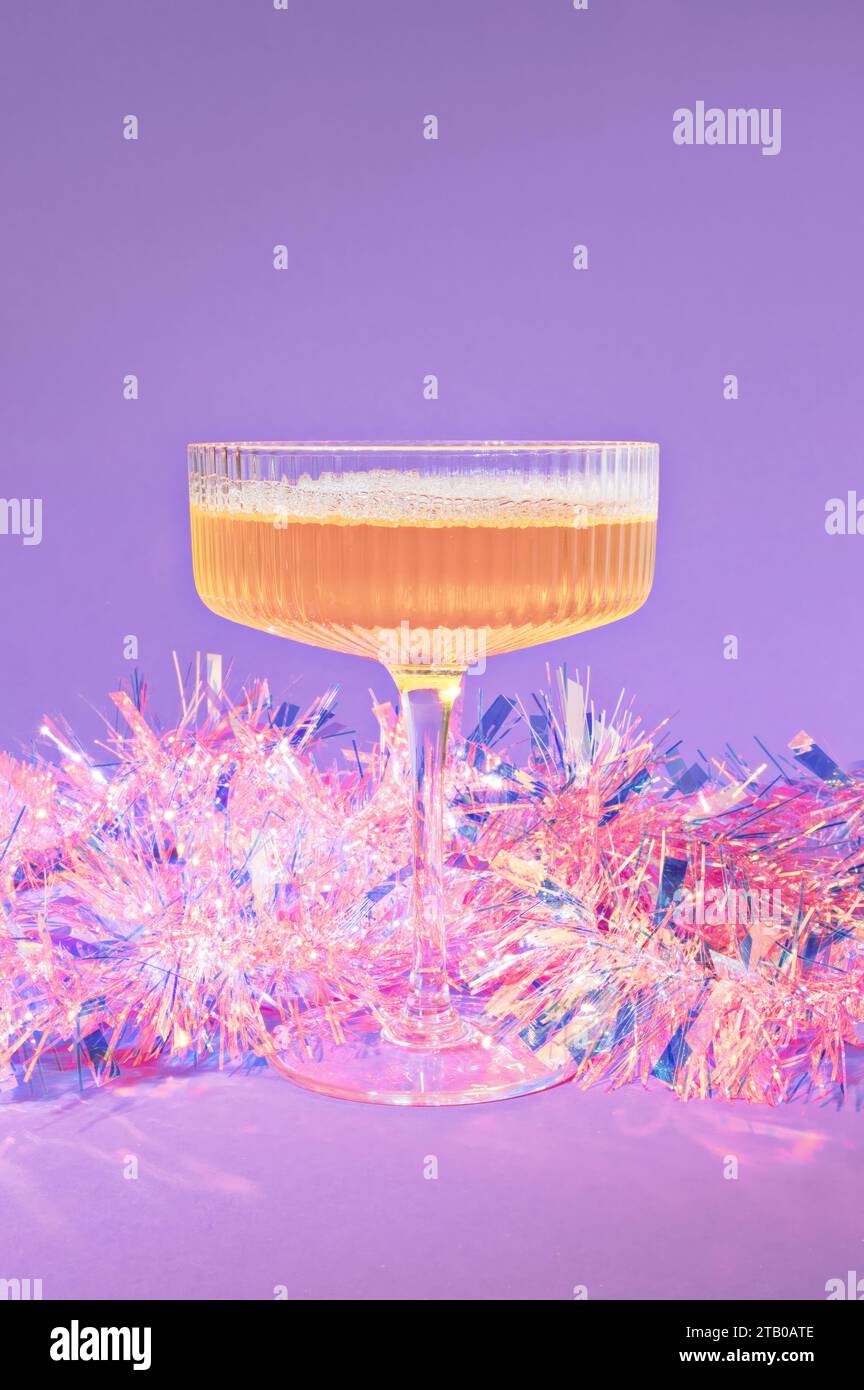 Ein Weihnachtscocktail mit Champagner, verpackt in rosa schillerndem Lametta-Weihnachtsdekoration auf lila Hintergrund Stockfoto