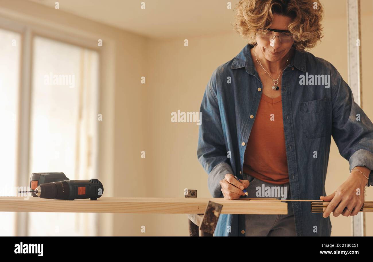 Reife weibliche Umbauerin, die an einem Küchen-Renovierungsprojekt arbeitet. Sie misst und markiert die Fußleisten mit einem Maßband und Bleistift. Karpfenqualifikation Stockfoto