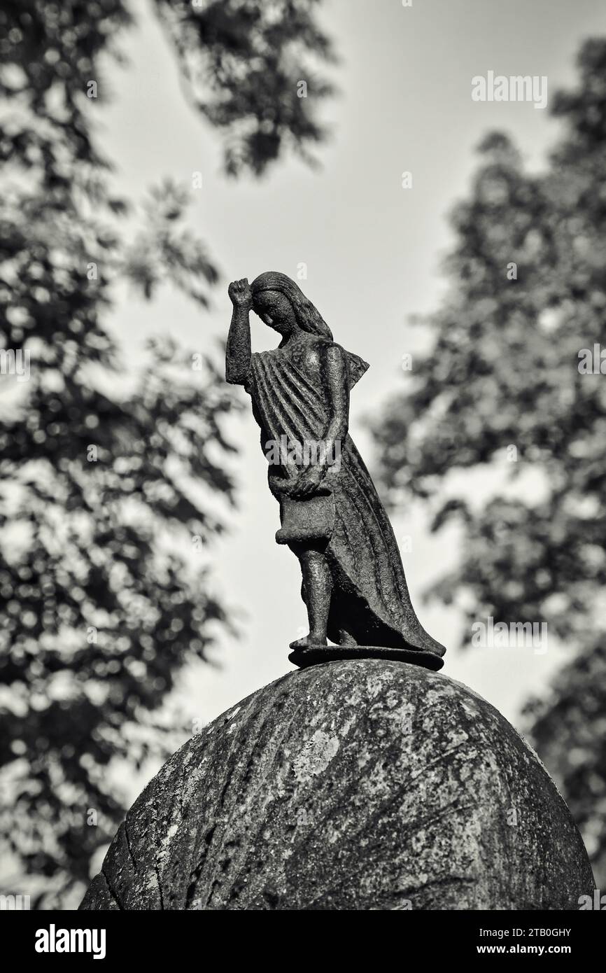 Traurige Botenfigur auf altem Grabstein, Bokeh-Hintergrund mit herbstlichem Laub. Schwarzweißkonvertierung. Sauvo Kirchfriedhof, Sauvo, Finnland. Stockfoto
