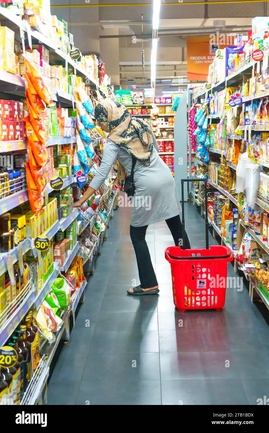 November 2023. Surabaya, Indonesien. Eine muslimische Frau im Hijab untersucht ein Regal mit bunten Produkten in einem hell beleuchteten Supermarktgang. Straße Stockfoto