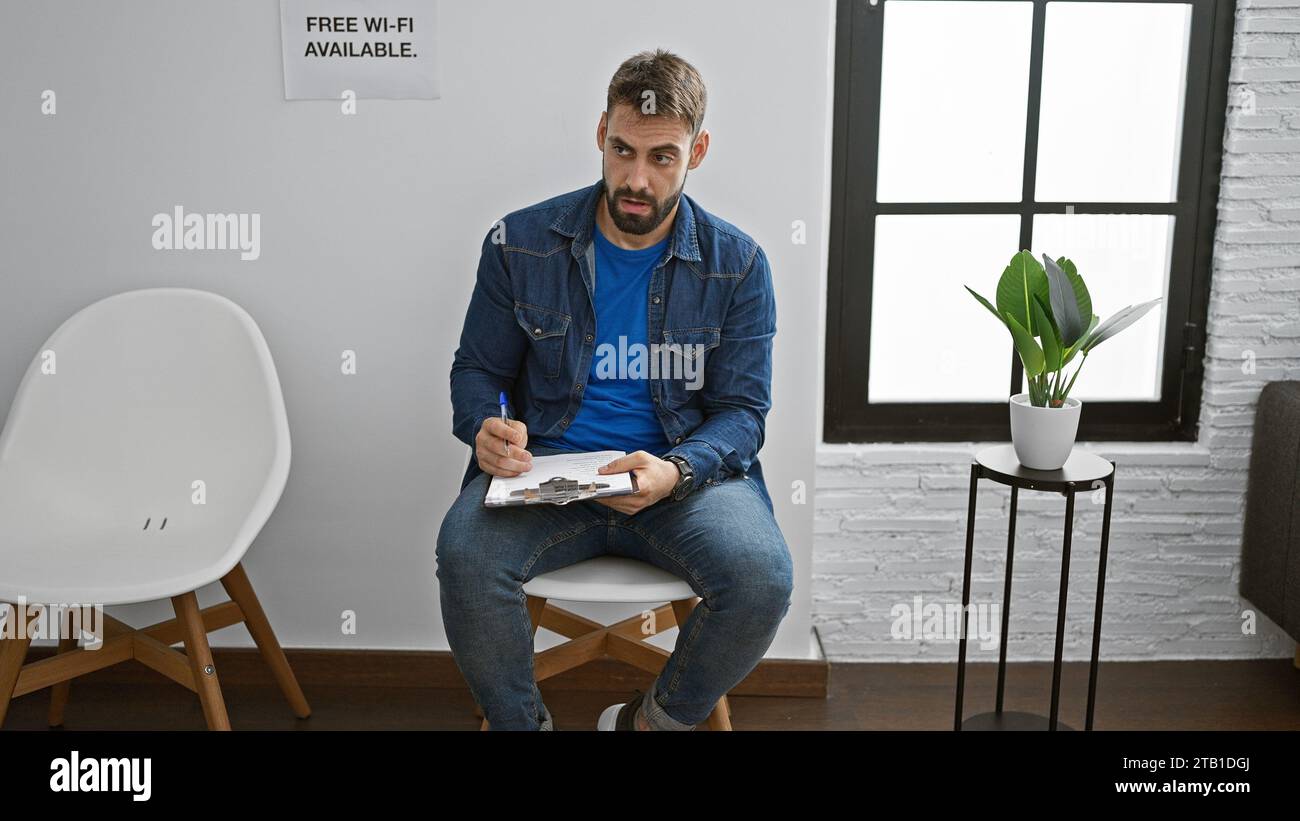 Der hübsche junge hispanische Mann, ein ernster Erwachsener mit Bart, entspannt sich in einem Stuhl, tief vertieft darin, einen Bericht auf einem Klemmbrett in einer ruhigen Inneneinrichtung zu schreiben Stockfoto