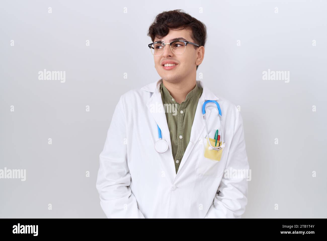 Junger, nicht binärer Mann, der Ärzteuniform und Stethoskop trägt und mit einem Lächeln im Gesicht zur Seite blickt, natürlicher Ausdruck. Lacht selbstbewusst. Stockfoto
