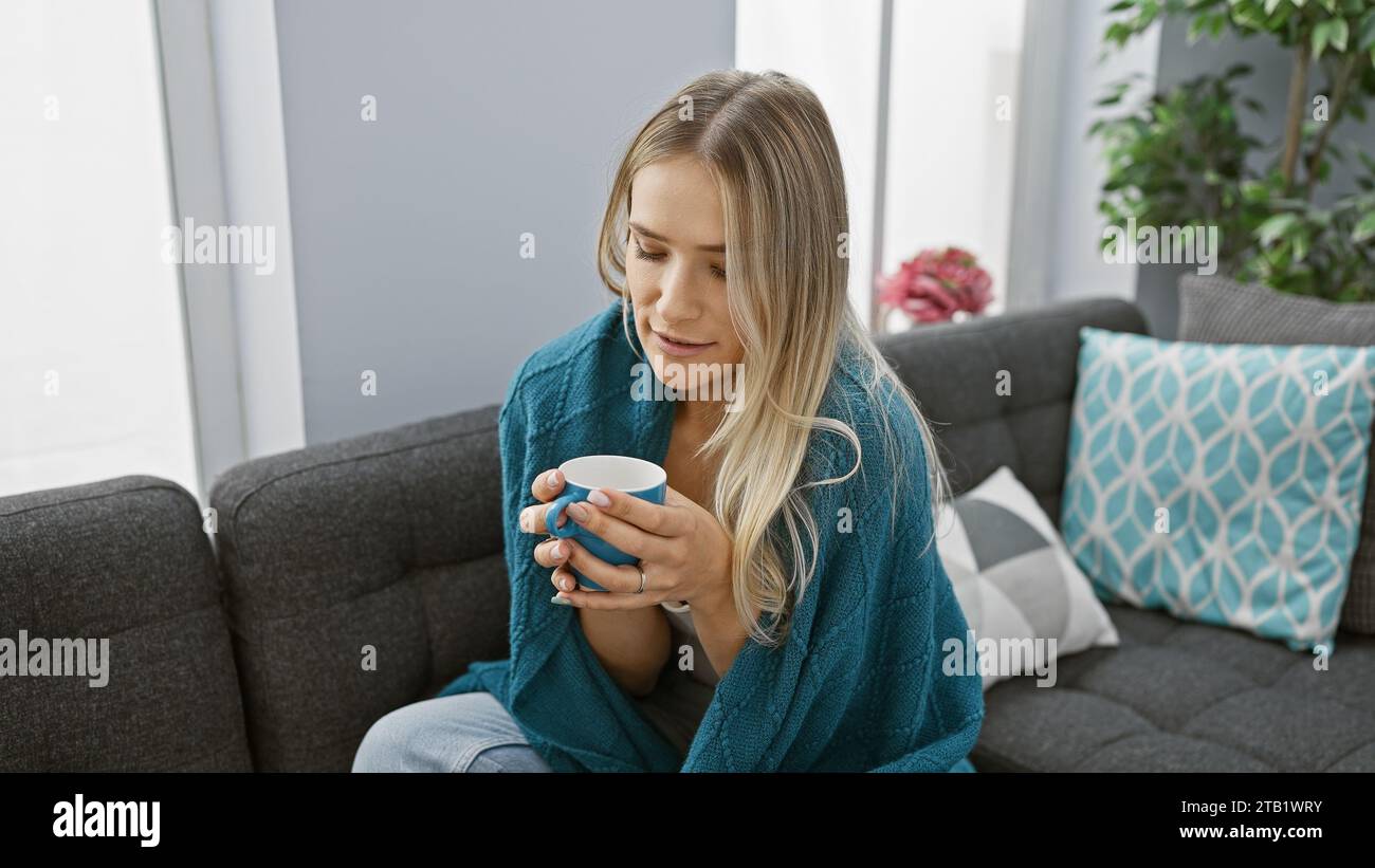 Schöne junge blonde Frau, die zu Hause in eine gemütliche Decke gewickelt ist und sich an einem kalten Wintertag mit einer heißen Tasse Kaffee aufwärmt Stockfoto Schöne junge blonde Frau, die zu Hause in eine gemütliche Decke gewickelt ist und sich an einem kalten Wintertag mit einer heißen Tasse Kaffee aufwärmt Stockfoto