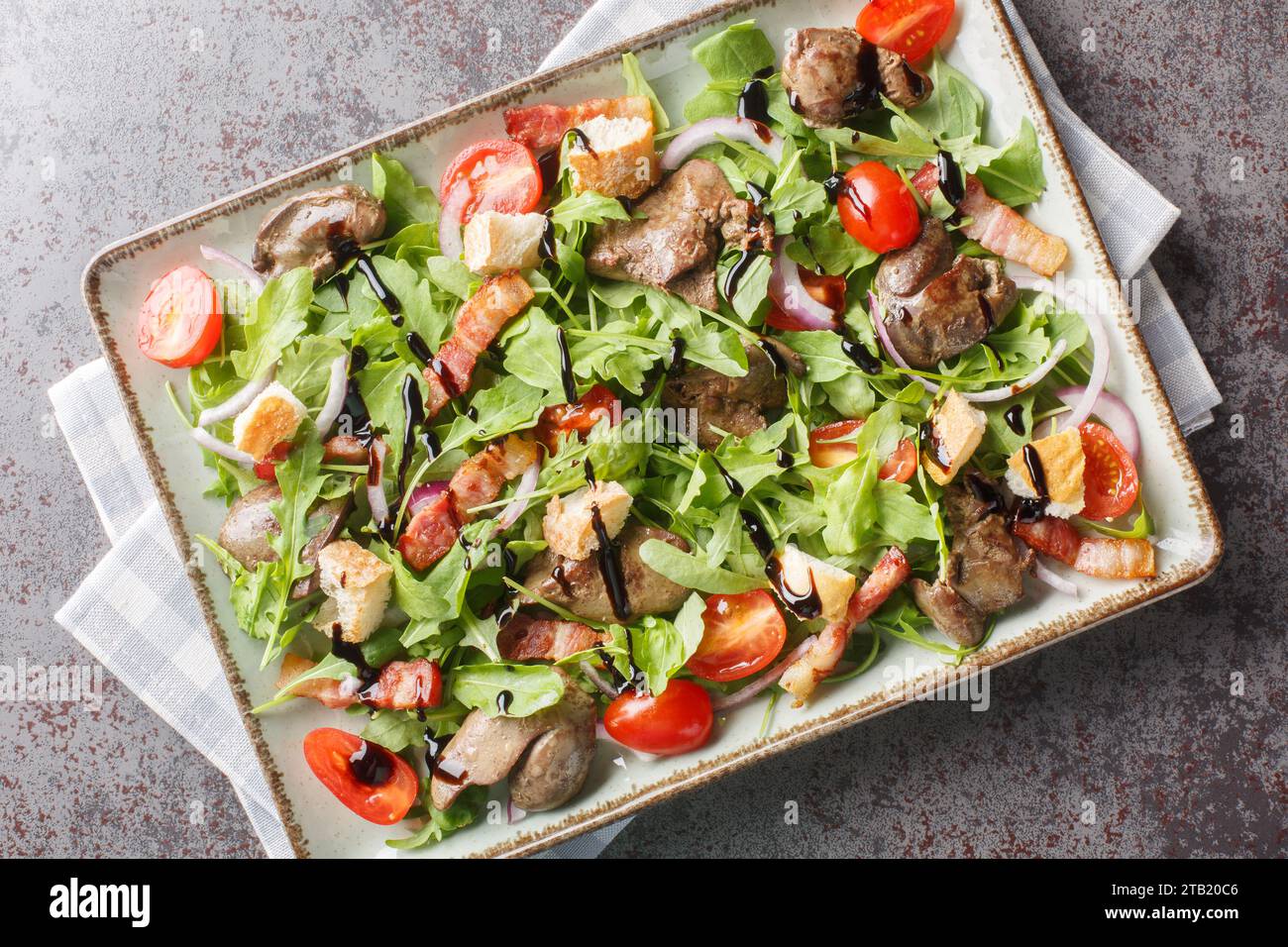 Köstlicher diätetischer Salat aus Hühnerleber, Rucola, Zwiebeln, Tomaten, Croutons und Speck in Nahaufnahme auf einem Teller auf dem Tisch. Horizontale Draufsicht von abo Stockfoto