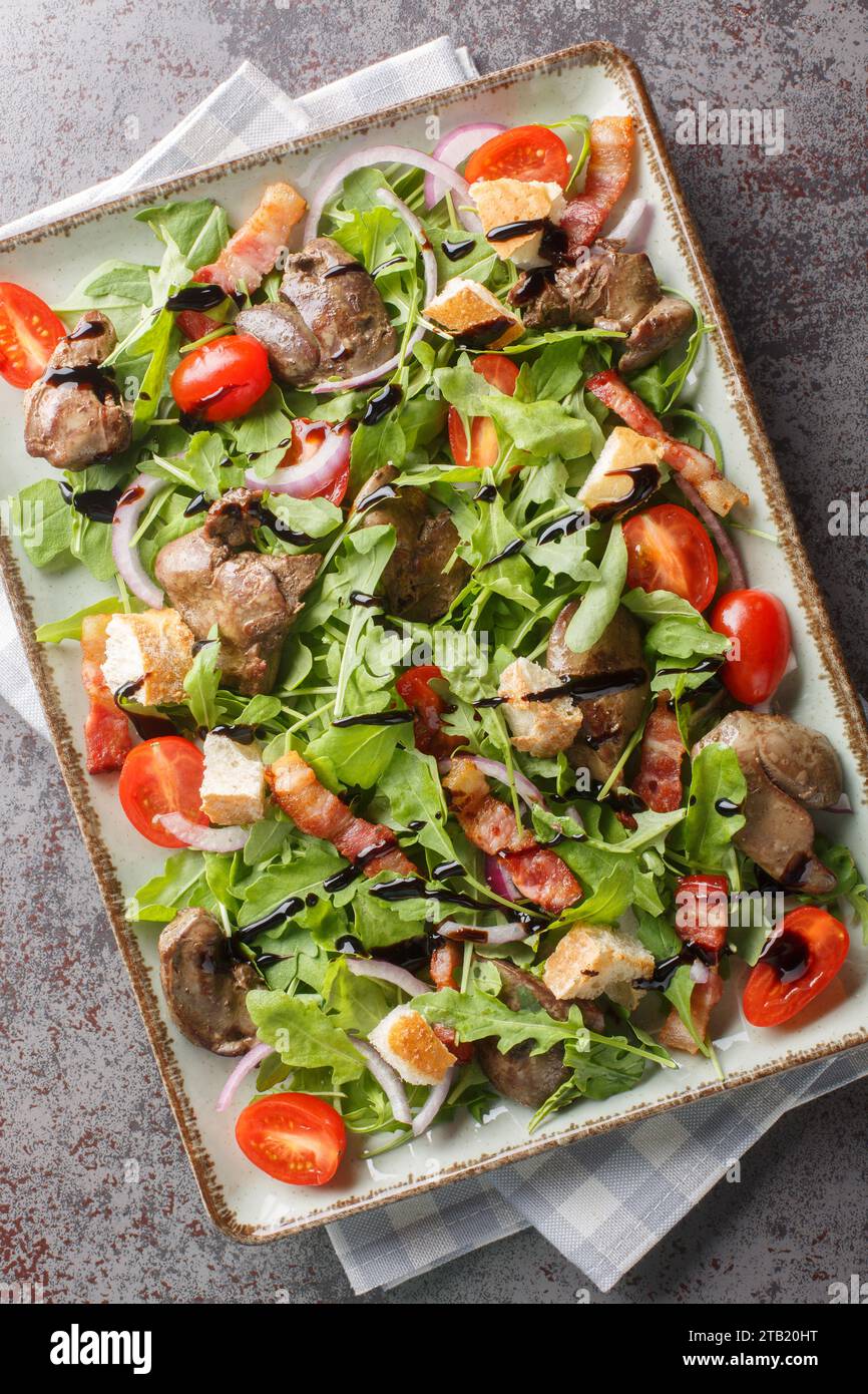 Warmer Hühnerlebersalat mit Speck, Kirschtomaten, Rucola. Rote Zwiebeln und Croutons in Nahaufnahme auf dem Teller auf dem Tisch. Vertikale Draufsicht von oben Stockfoto