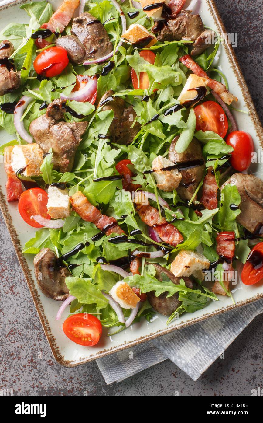 Warmer Salat mit gegrillter Hähnchenleber, Speck, Kirschtomaten, Rucola, roten Zwiebeln und Croutons in Nahaufnahme auf dem Teller auf dem Tisch. Vertikale Draufsicht von abo Stockfoto