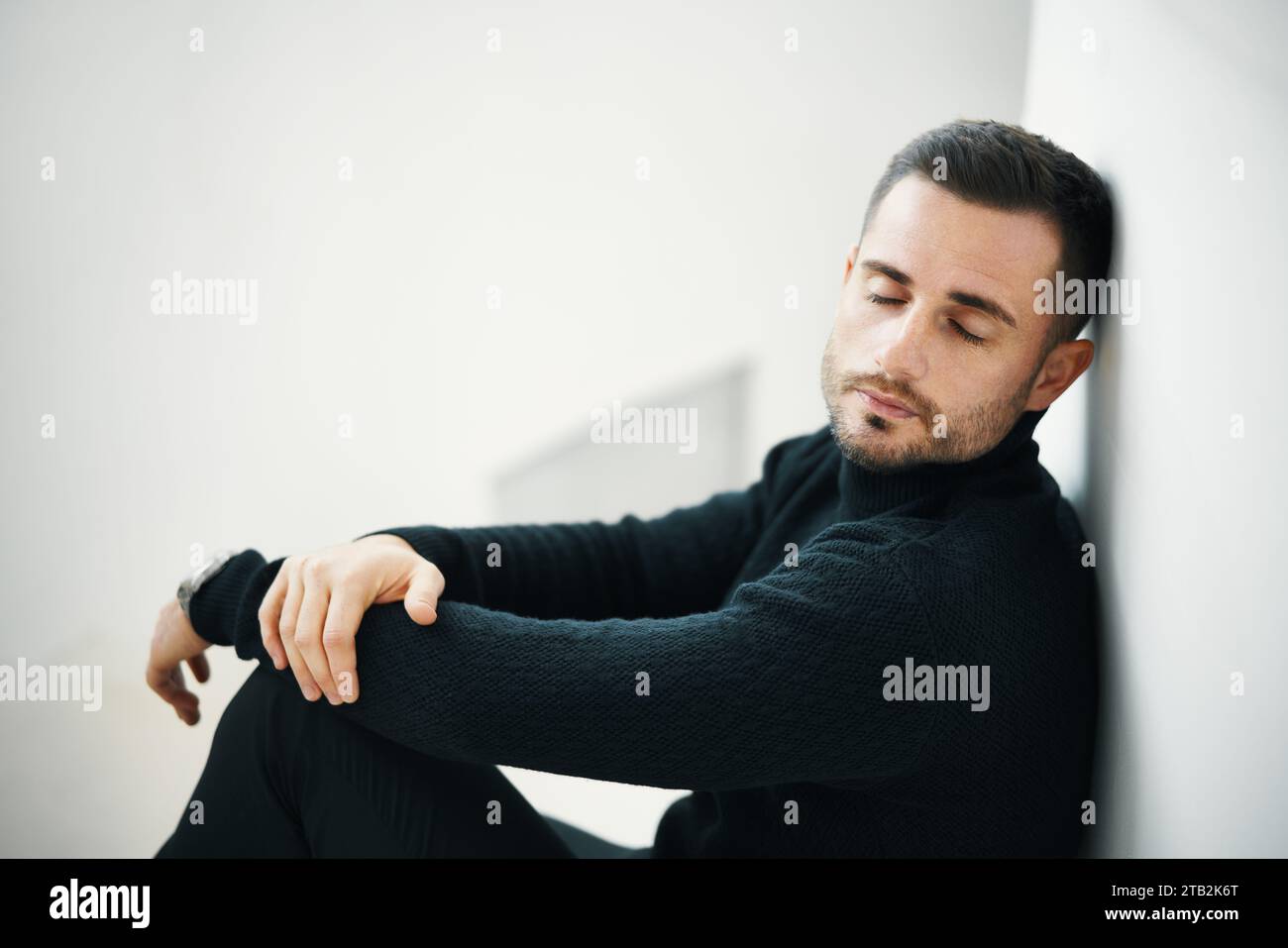 Müder Mann in einem schwarzen Pullover ruht mit geschlossenen Augen an einer weißen Wand. Relax-Konzept Stockfoto