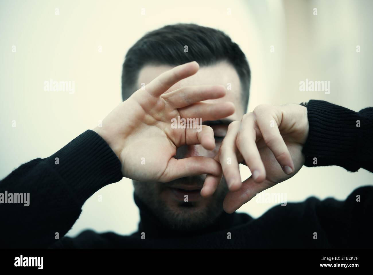 Ein junger Mann mit einem dunklen Rollkragen bedeckt sein Gesicht mit den Händen in einem nachdenklichen oder emotionalen Moment. Das Konzept der Emotionen von Menschen Stockfoto