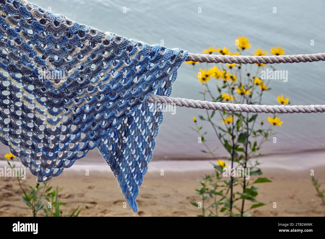 Eine Flechtdecke aus Spitze in blauer Farbe, die an den Seilen vor dem Hintergrund von Blumen und einem Sandstrand vergessen wurde. Stockfoto