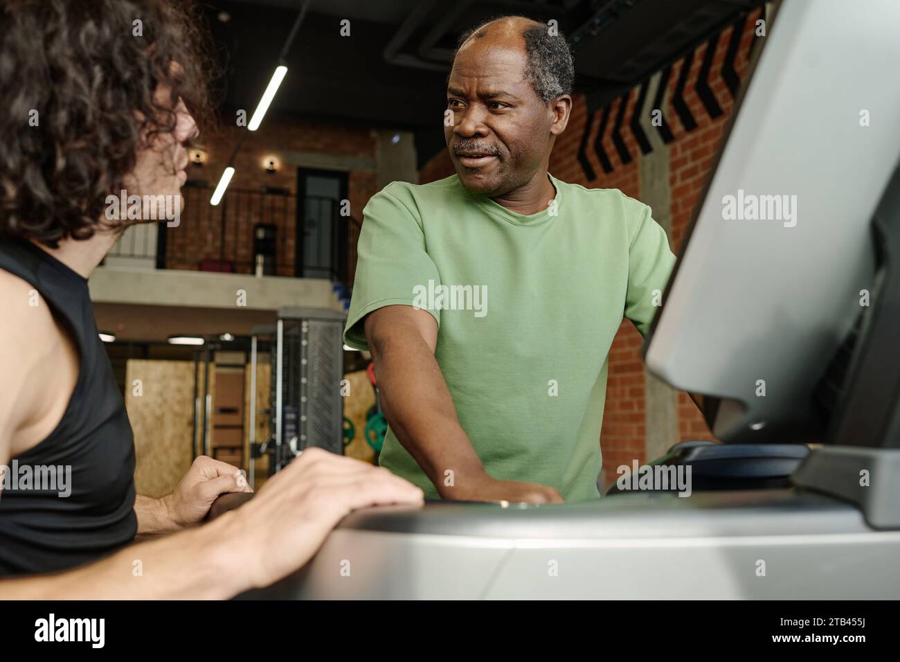 Ein älterer schwarzer Mann, der im Fitnessstudio auf dem Laufband steht und mit dem jungen kaukasier neben ihm spricht Stockfoto