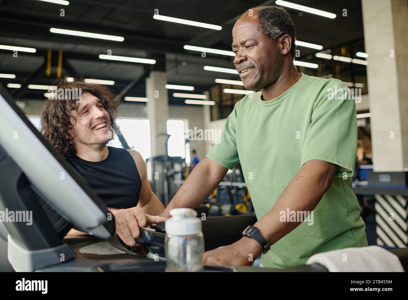 Ein älterer schwarzer Mann, der im Fitnessstudio auf dem Laufband läuft, mit einem jungen kaukasischen Athleten neben ihm Stockfoto