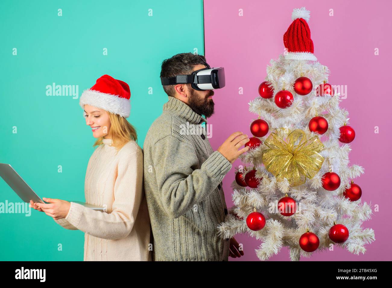 Bärtiger Mann im VR-Headset und hübsches Mädchen im Weihnachtsmann-Hut mit Laptop in der Nähe des Weihnachtsbaums. Moderne Technologie und Unterhaltung. Weihnachtsspaare spielen Stockfoto