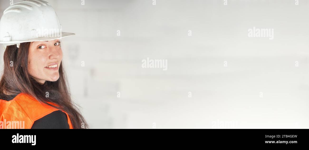 Junge Frau mit weißem Schutzhelm und orangefarbener Warnweste, langes dunkles Haar, schaut über ihre Schulter in die Kamera, lächelt. Unscharfe Konstruktion si Stockfoto