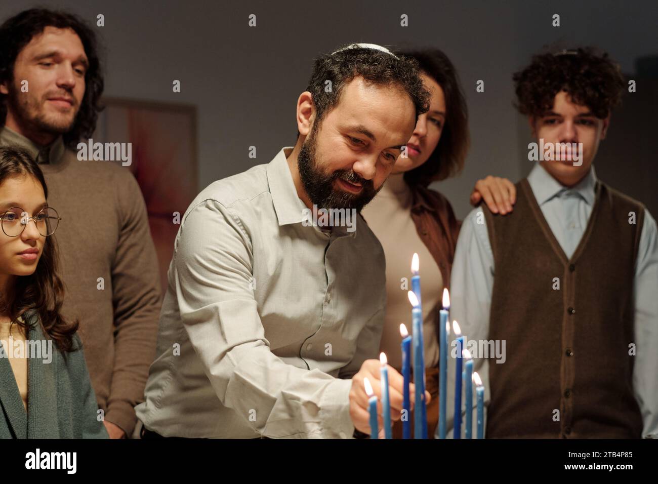 Reifer Mann in jüdischem Schädelkappen, der brennende blaue Kerze auf den Leuchter der Menora setzt, während er Hanukka zu Hause mit seiner Familie feiert Stockfoto